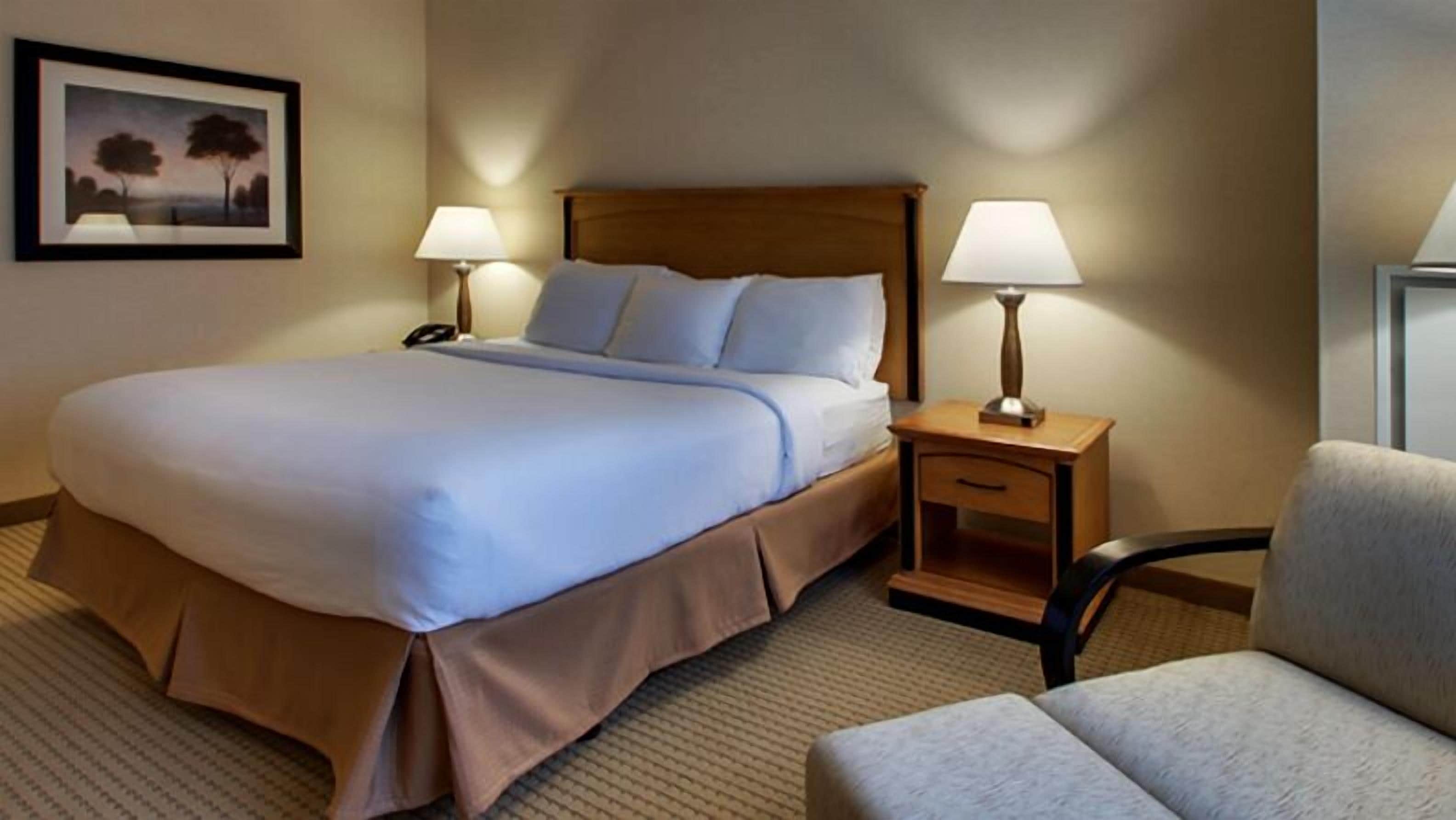 Een hotelkamer met een groot bed, twee nachtkastjes met lampen, een stoel en een inlijsting foto aan de muur.