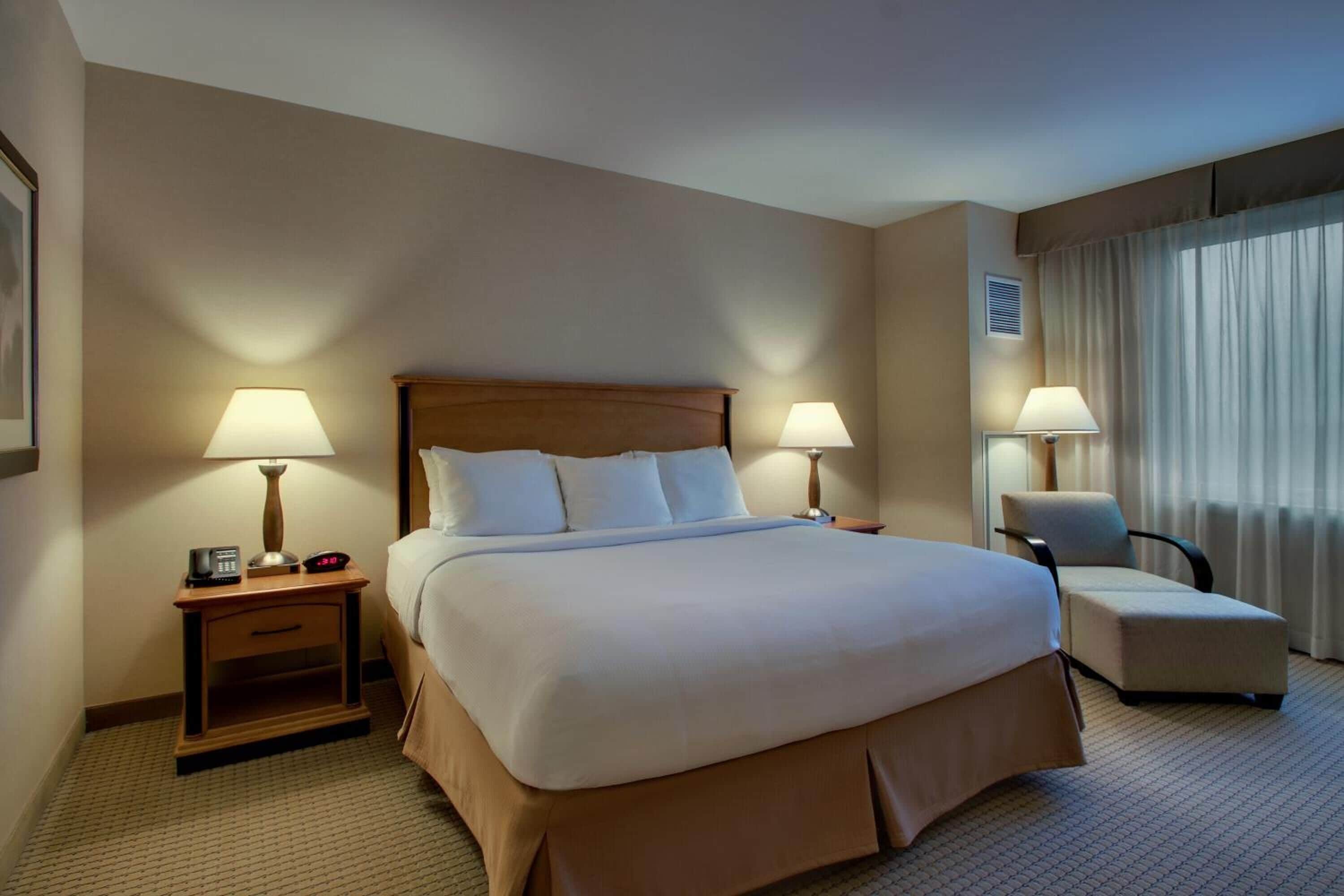 A hotel room with a large bed, two bedside tables with lamps, a chair, and a window with curtains.