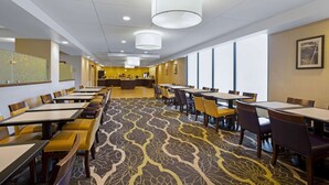 Une salle à manger d’hôtel avec plusieurs tables et chaises, un tapis à motifs et un comptoir de réception en arrière-plan.