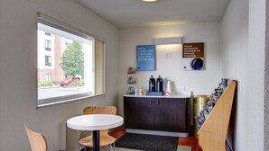 A small hotel room with a window, a desk with a chair, a cabinet, and a coffee machine.
