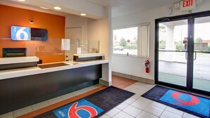 A hotel reception area with a digital sign displaying room number 6, a reception desk, a fire extinguisher, and a glass door leading outside.
