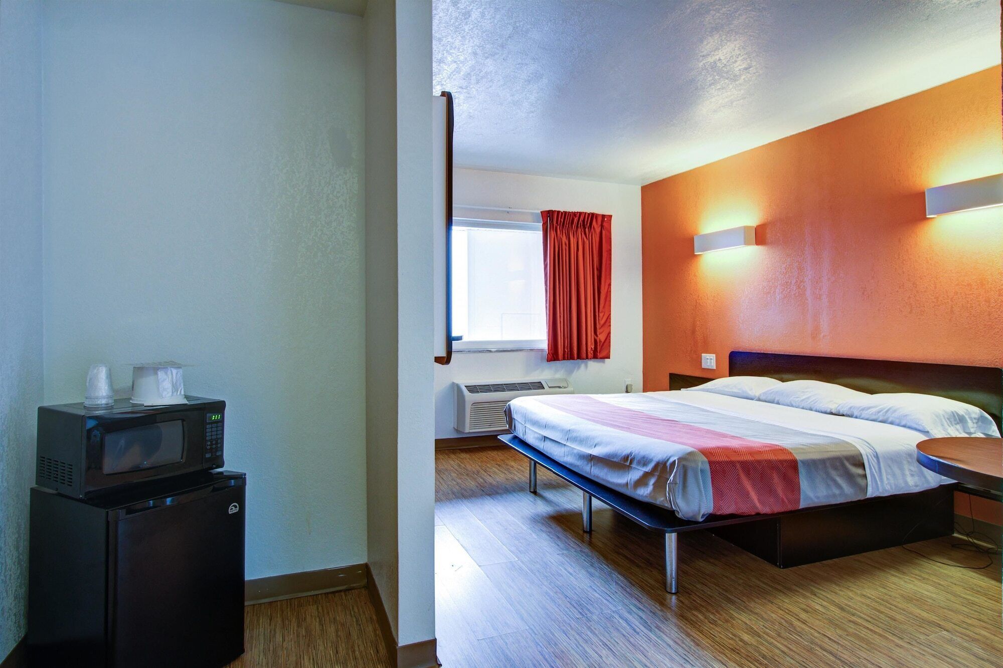 Une chambre d’hôtel comprenant un lit, un bureau avec un micro-ondes, une fenêtre avec des rideaux et un lampadaire fixé au mur.