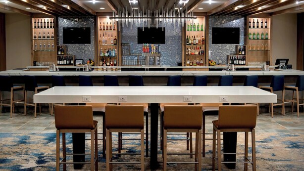 A modern bar area with a long counter, multiple seating options, and a variety of liquor bottles. - Delta Hotels by Marriott Utica (Utica)