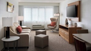 A hotel room with a sofa, ottoman, armchair, TV, lamp, and a small table. - Delta Hotels by Marriott Utica (Utica)