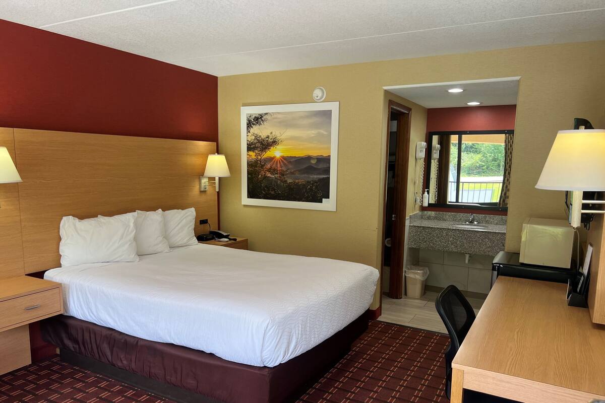 A hotel room with a large bed, bedside lamps, a desk with a chair, a painting of a sunset, and a bathroom visible through an open door.
