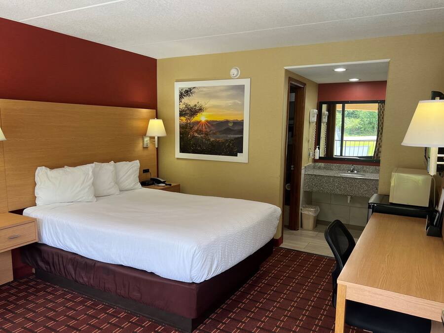 A hotel room with a large bed, bedside lamps, a desk with a chair, a painting of a sunset, and a bathroom visible through an open door.