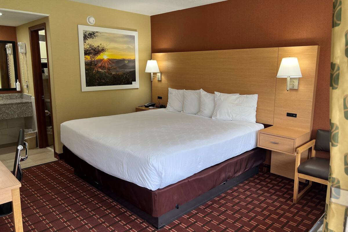 A hotel room with a large bed, two bedside tables, a nightstand, a headboard, a painting, a mirror, a telephone, a lamp, a chair, and a carpeted floor.