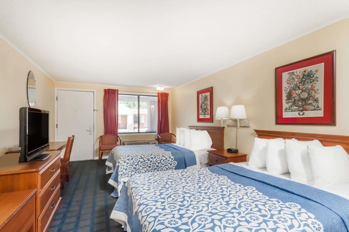 A hotel room with two beds, a desk with a television, a chair, a window with red curtains, and framed artwork on the walls.