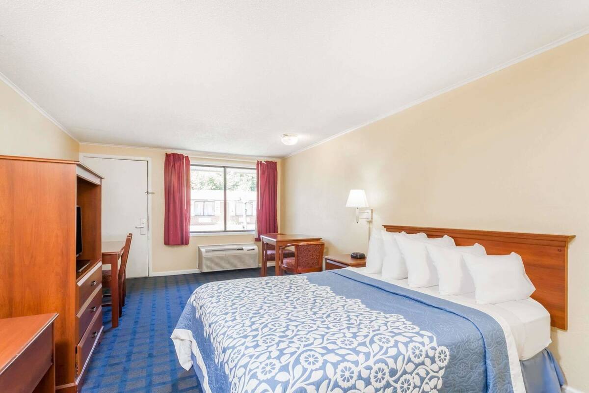 A hotel room with a bed, a desk, a chair, a TV, a window with curtains, and a lamp.