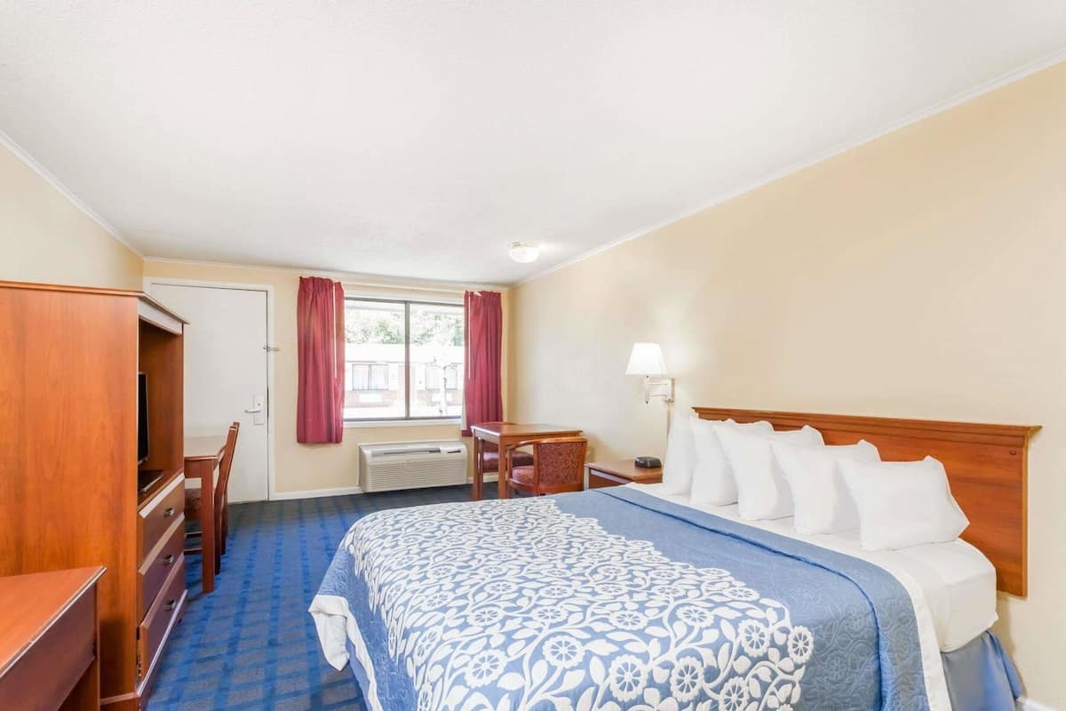 A hotel room with a bed, a desk, a chair, a TV, a window with curtains, and a lamp.