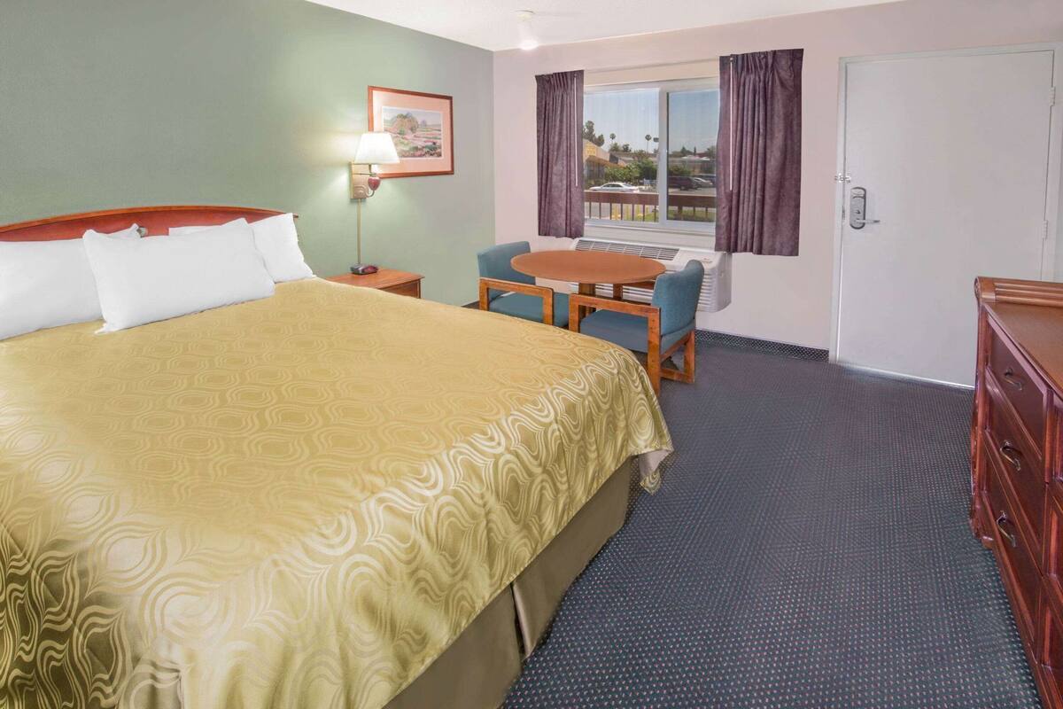 A hotel room with a bed, a desk, a chair, a lamp, a window with curtains, and a door.