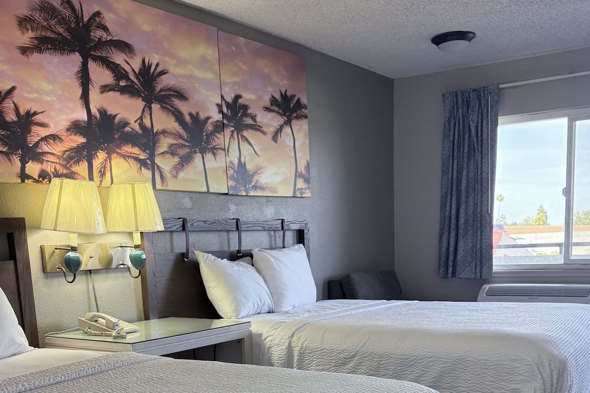 A hotel room with two beds, a nightstand with a lamp, a painting of palm trees, and a window with a view.
