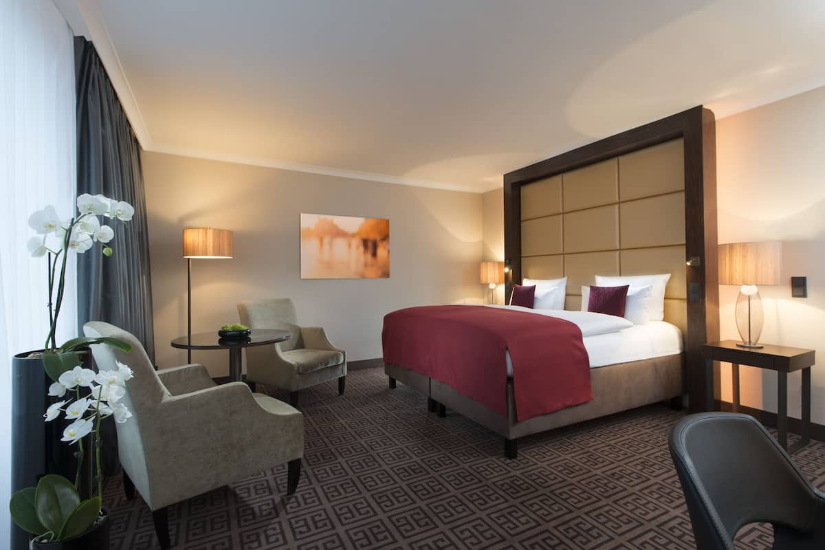 A hotel room with a large bed, two armchairs, a small table, a potted orchid, and a painting on the wall.