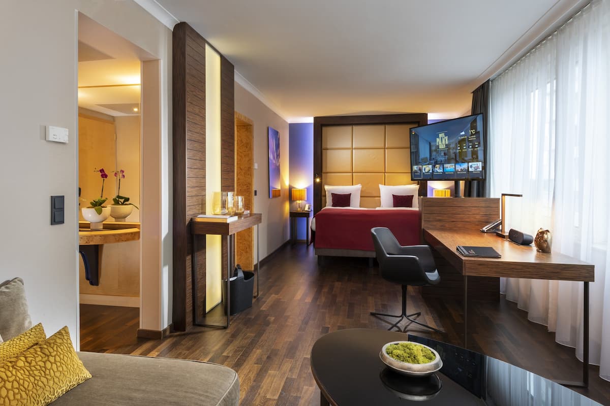 A modern hotel room with a bed, a desk, a TV, and a seating area.
