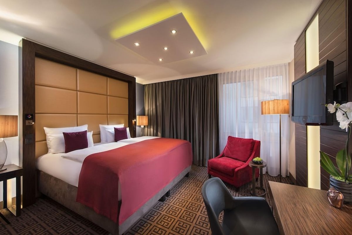 A hotel room with a large bed, a red chair, a desk, a television, and a plant.