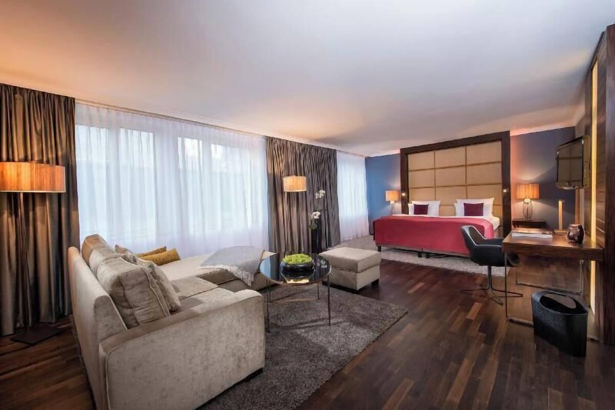 A modern hotel room with a sofa, ottoman, and a desk with a lamp.