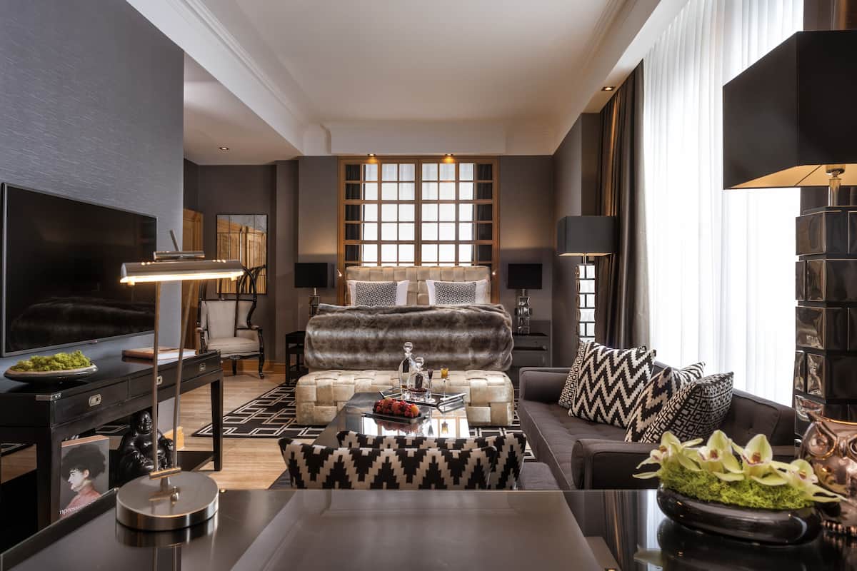 A modern living room with a sofa, coffee table, and TV. There is a large window with blinds, a fireplace, and a bookshelf.