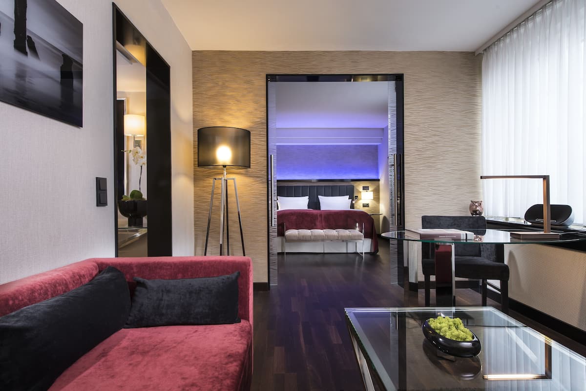 A modern hotel room with a red sofa, a glass coffee table, a desk with a lamp, and a view of the city through a large window.