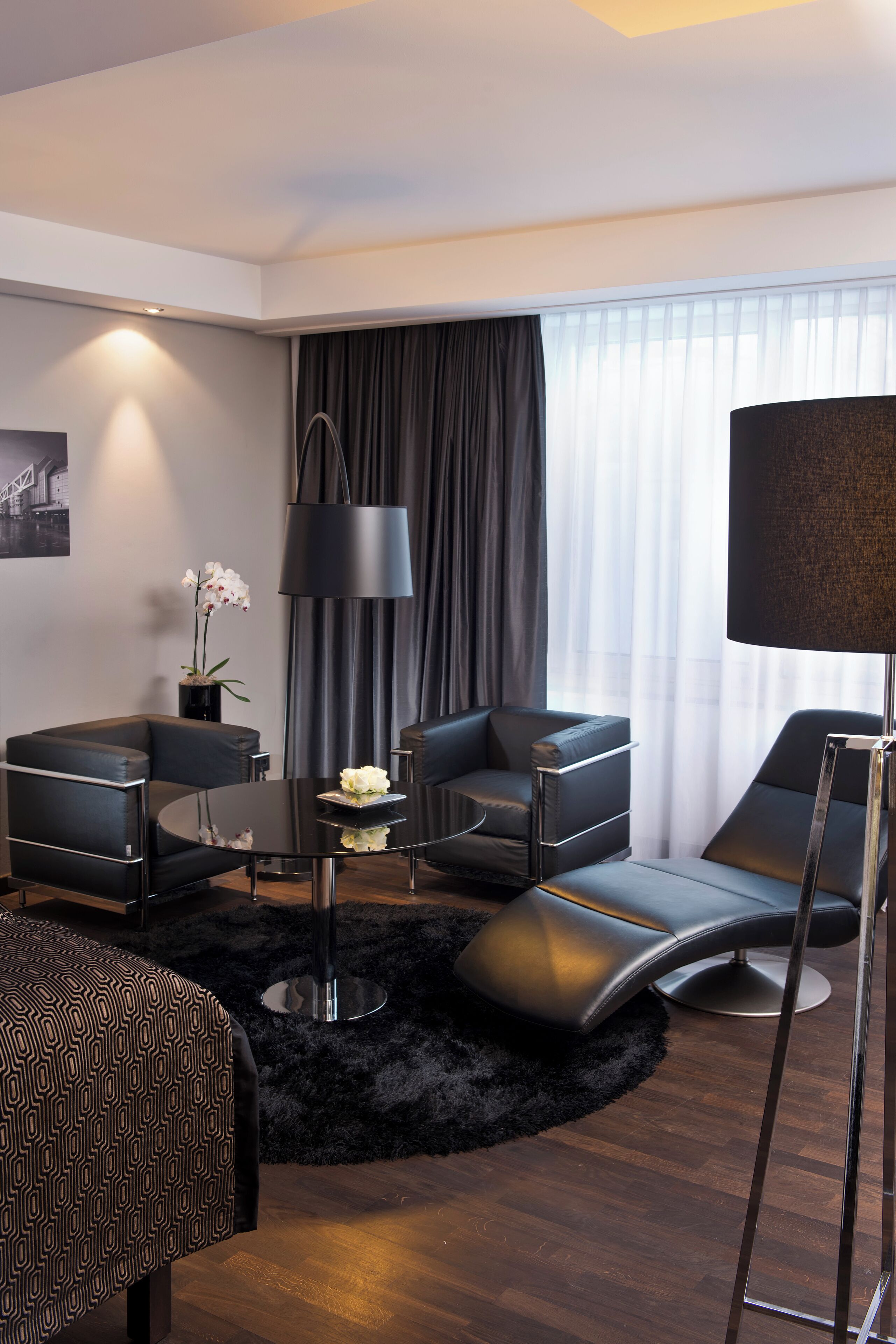 Habitació d'hotel moderna amb un sofà de pell negra, una taula de cafè rodona i un armari de seients negre. Hi ha una catifa negra al terra, una làmpada negra i un vas amb flors blanques sobre la taula de cafè.