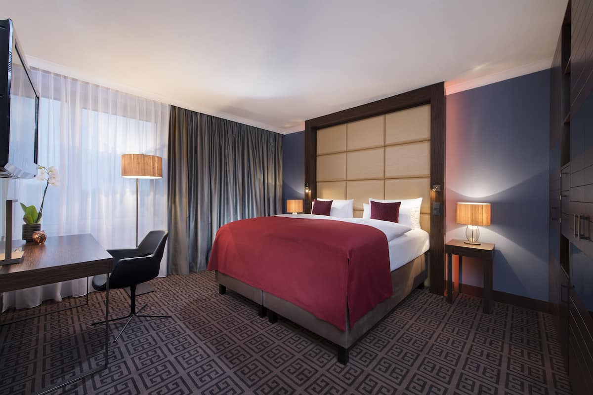A hotel room with a large bed, a desk with a chair, a TV, and a lamp.