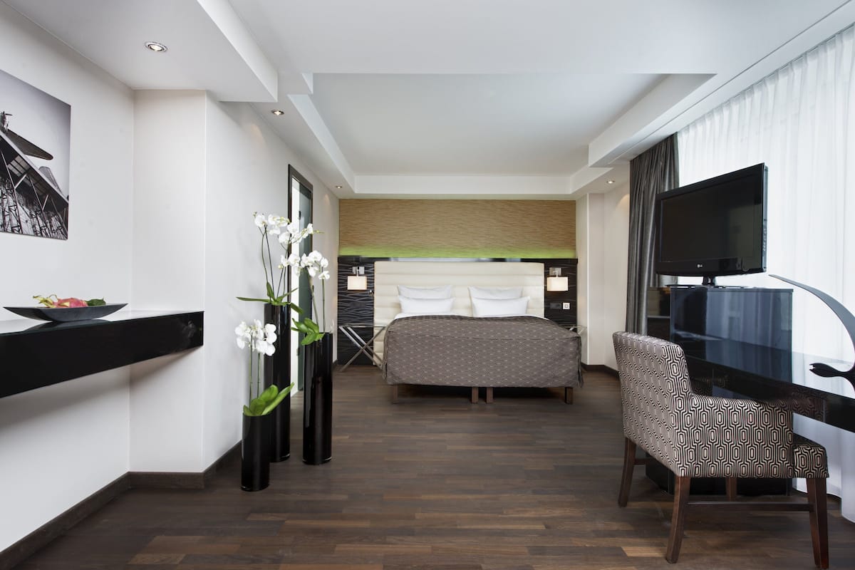 A hotel room with a bed, a desk with a TV, a chair, and a vase with flowers.