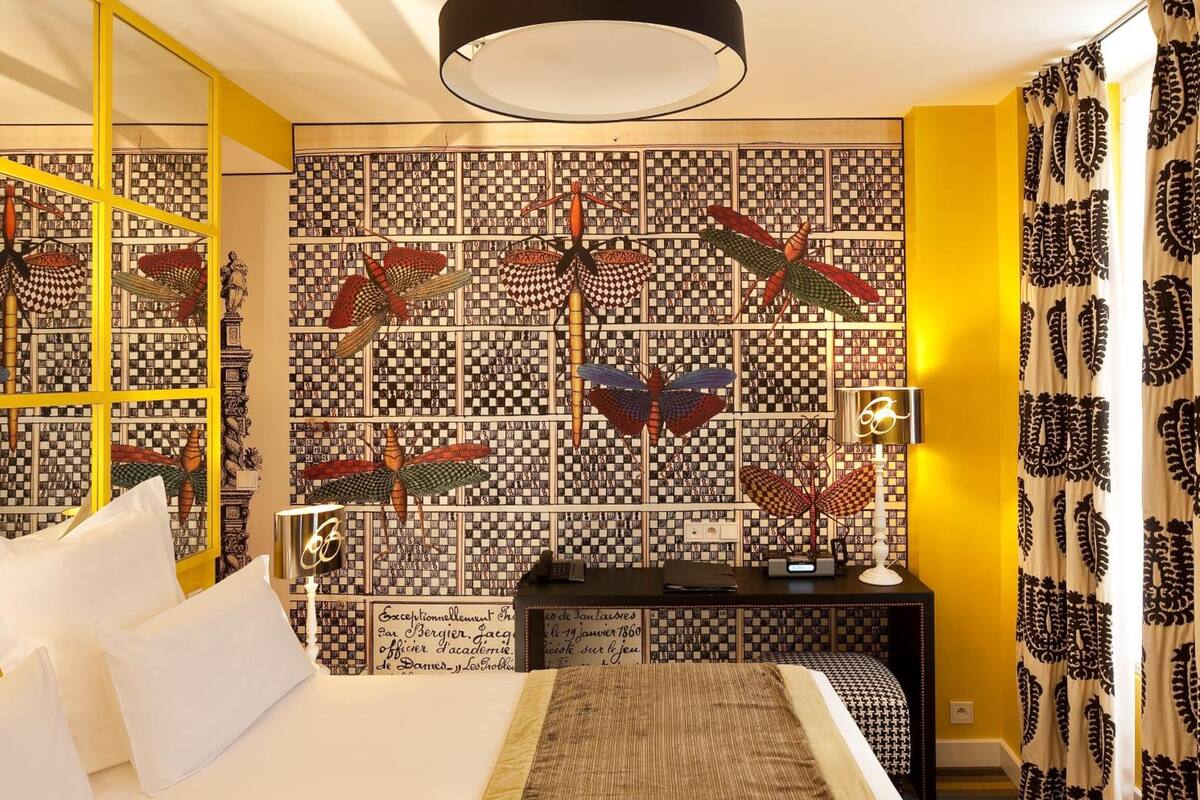 A bedroom with a checkered wallpaper, a bed with white linens, a nightstand with a lamp, and a mirror.