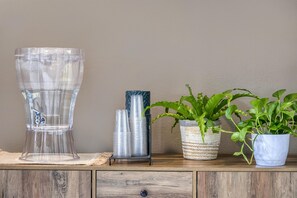 A clear glass punch bowl, a metal stack of plastic cups, and two potted plants on a wooden surface.