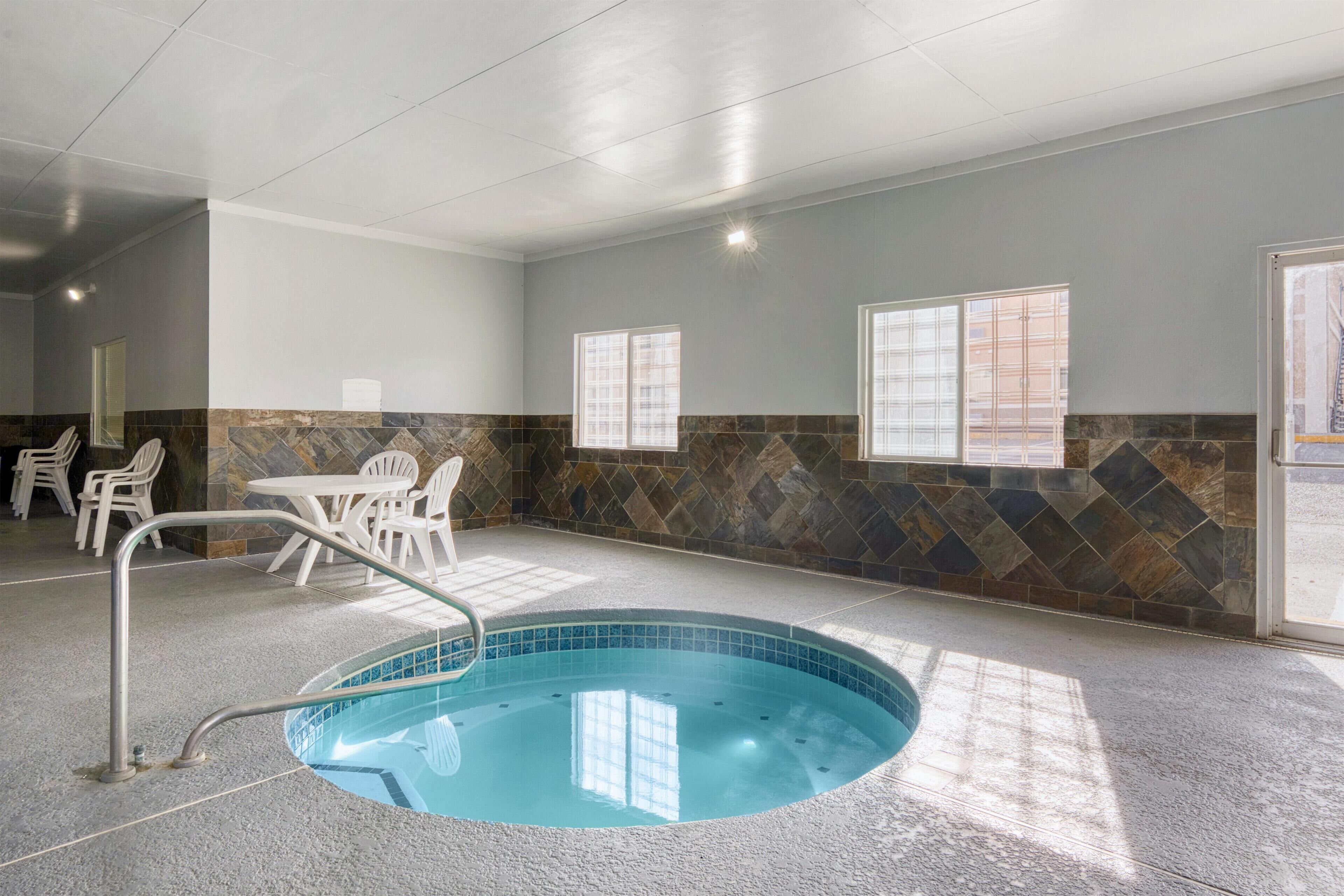 A swimming pool with a tiled edge, surrounded by a stone wall and a tiled floor. There are chairs and a table in the background.