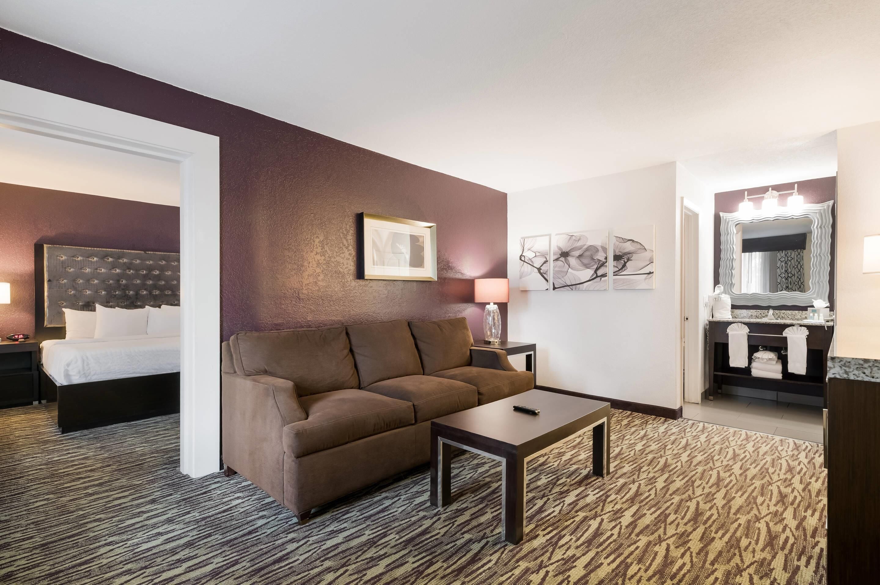 Hotelkamer met een bed, een bruine bank, een tafeltje, een lamp en een badkamer met spiegel en wastafel.