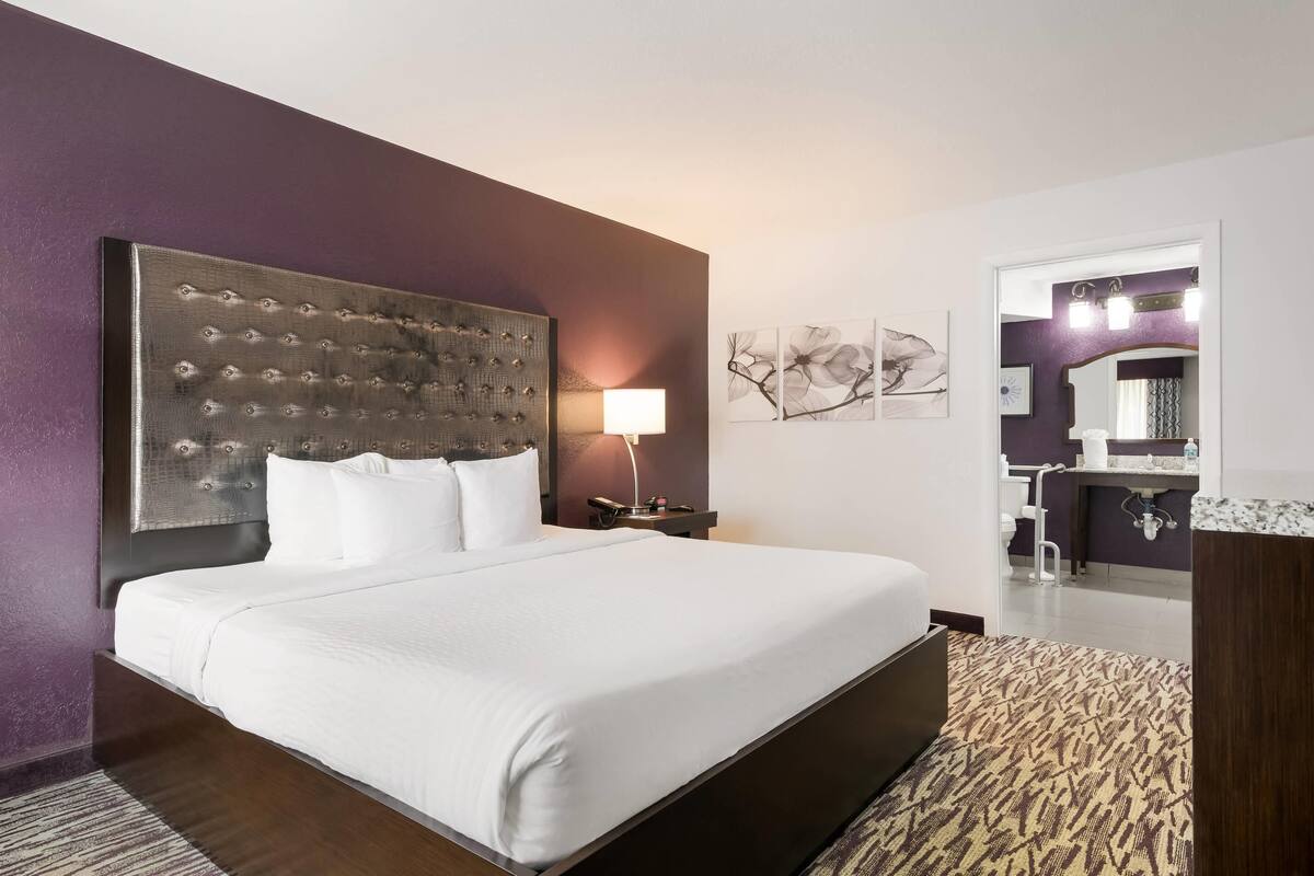 A hotel room with a large bed, a headboard, a bedside table with a lamp, a mirror, and artwork on the wall.