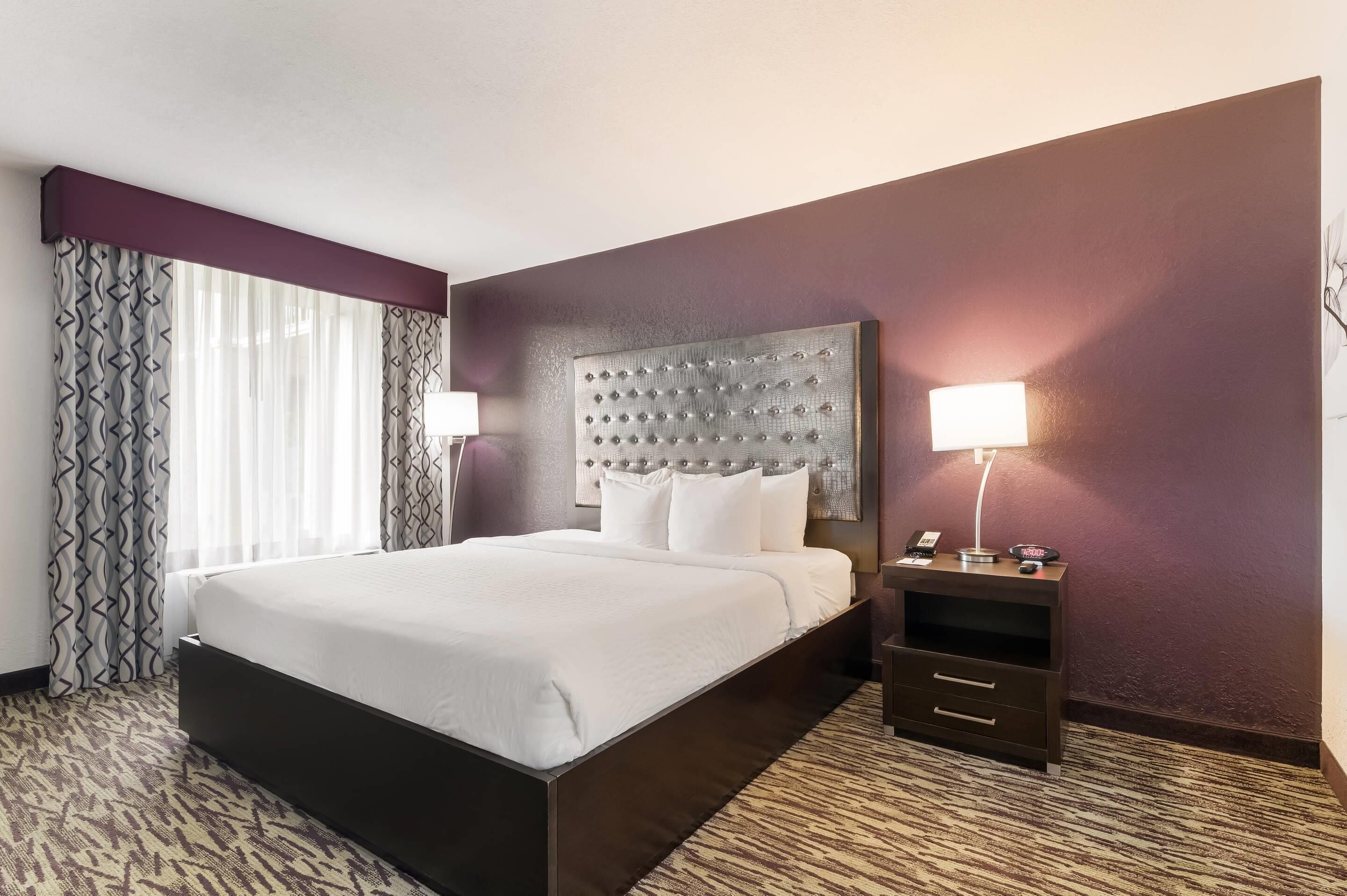 A hotel room with a large bed, a headboard with a tufted design, a bedside table with a lamp, and a small drawer unit.