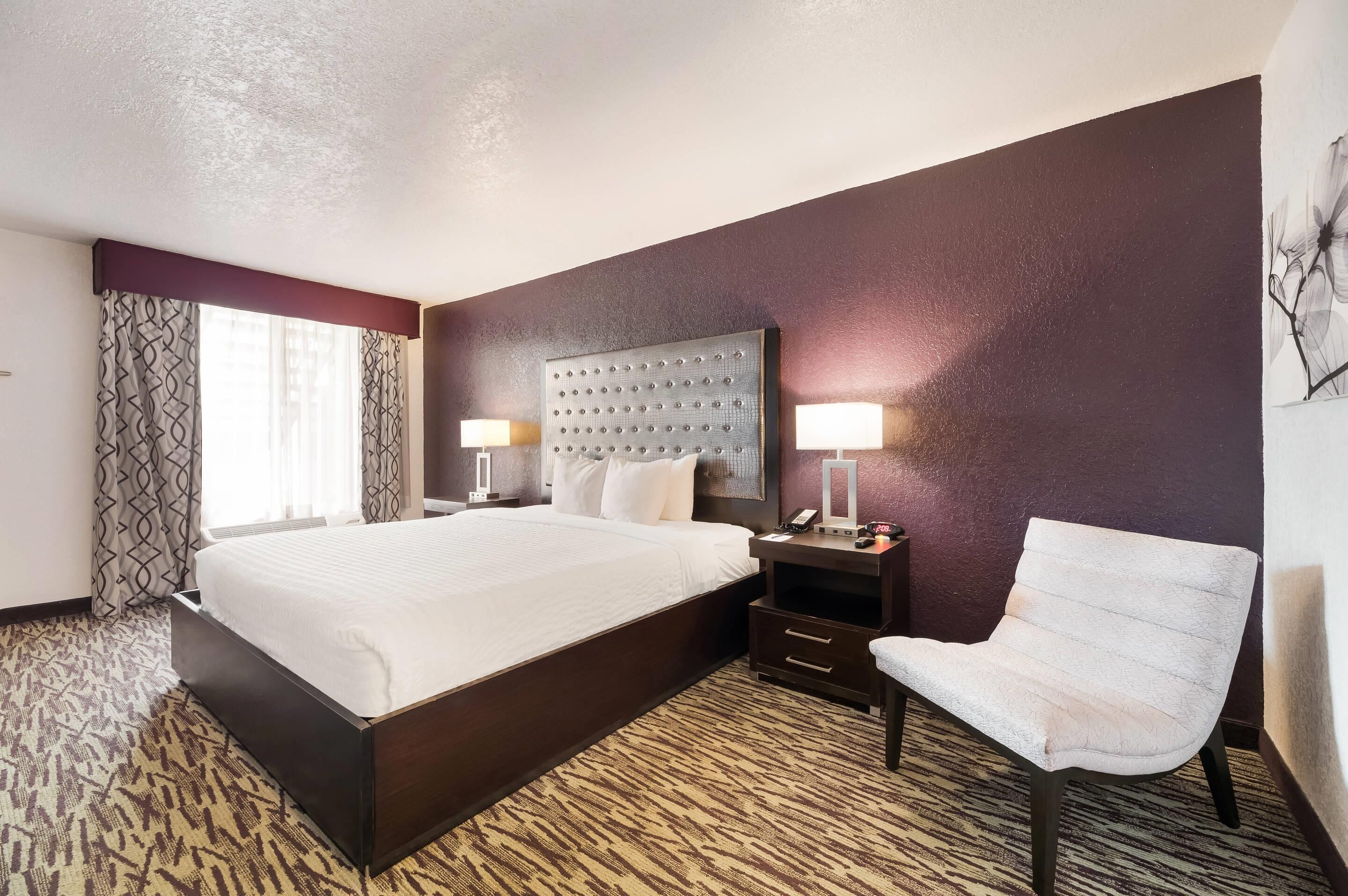 Hotelkamer met een groot bed, nachtkastje met lamp, stoel en een kunstwerk aan de muur.