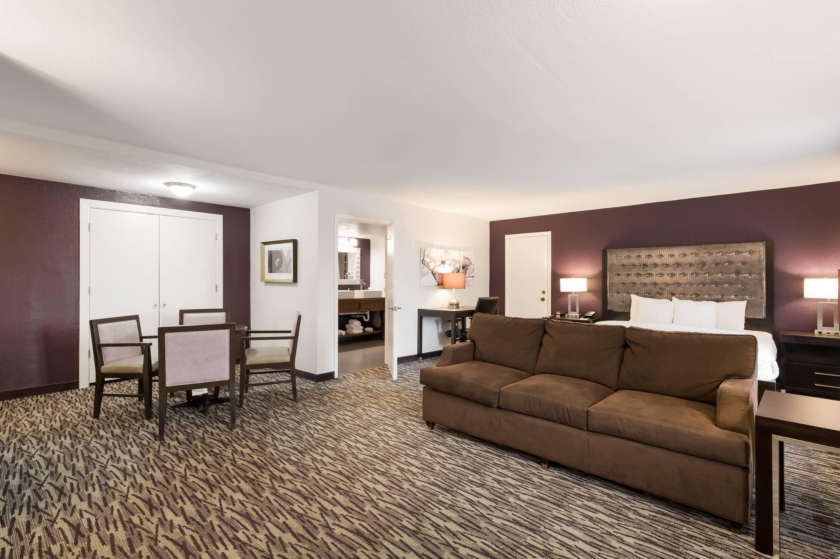 A hotel room with a brown sofa, a dining table with chairs, a small desk, and a lamp.