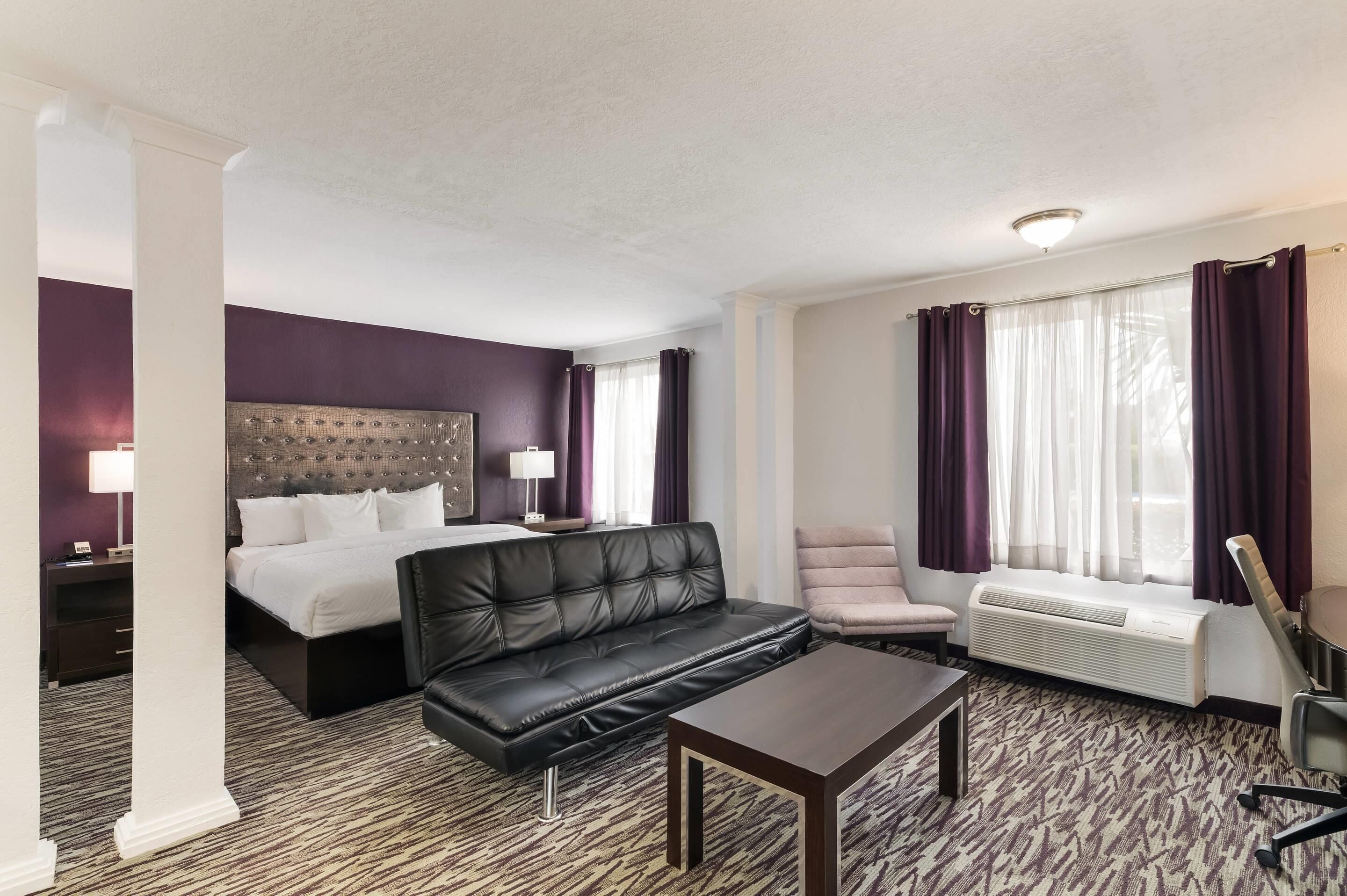 A hotel room with a bed, a black leather sofa, a small table, a chair, and a window with curtains.