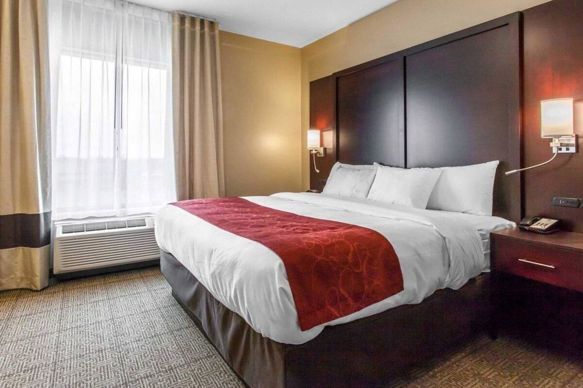A hotel room with a bed, bedside table, lamp, and an air conditioning unit.
