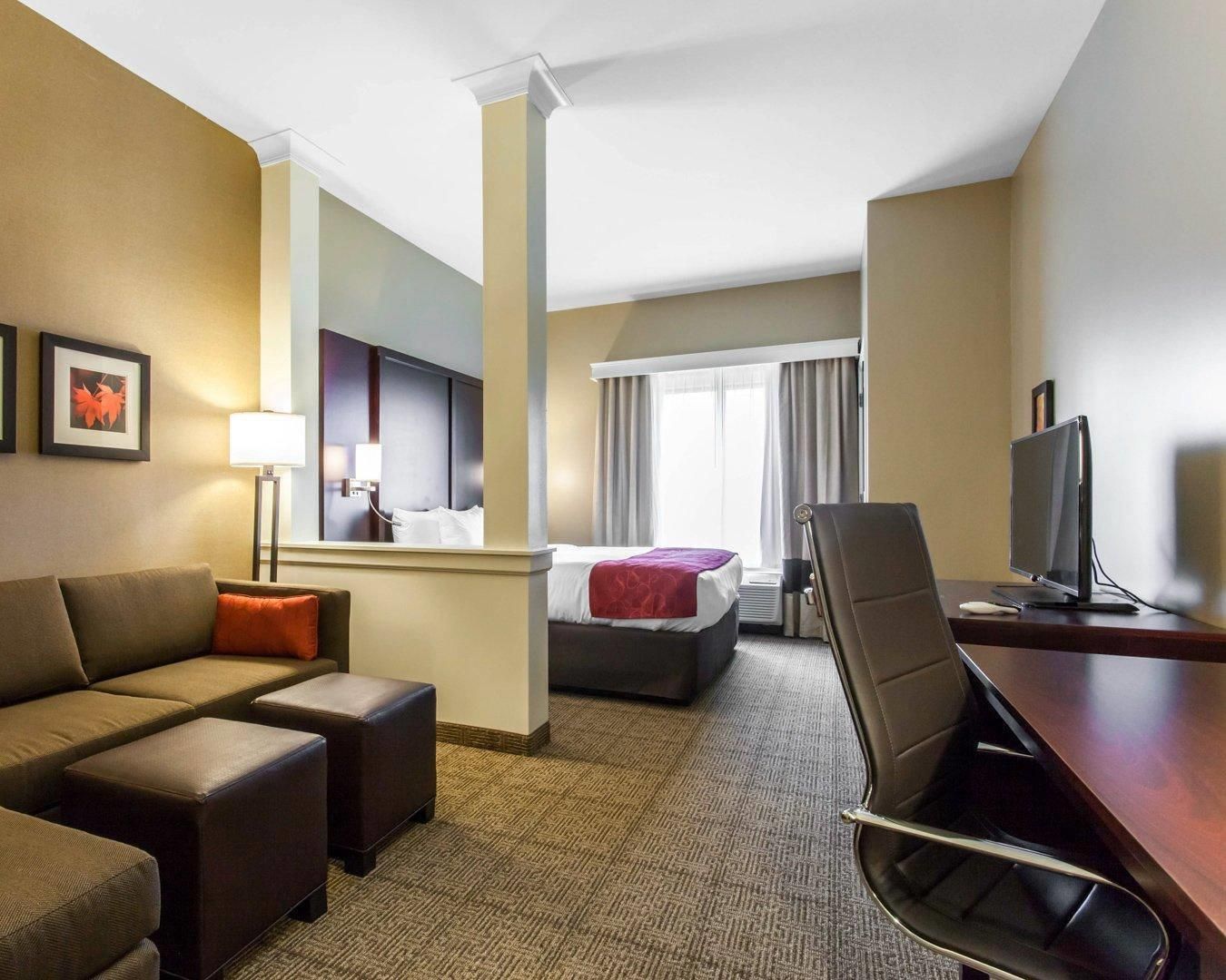 Ein Hotelzimmer mit Sofa, Schreibtisch mit Computer, Bett mit roter Decke, Fernseher und Fenster mit Vorhängen.