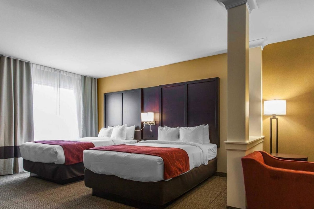 A hotel room with two beds, a nightstand, a lamp, and a chair.