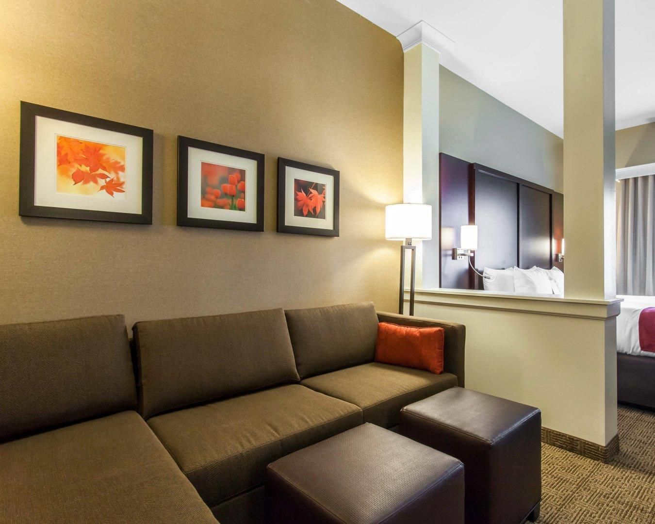 A hotel room with a sofa, ottoman, framed artwork, a lamp, and a bed with a headboard.