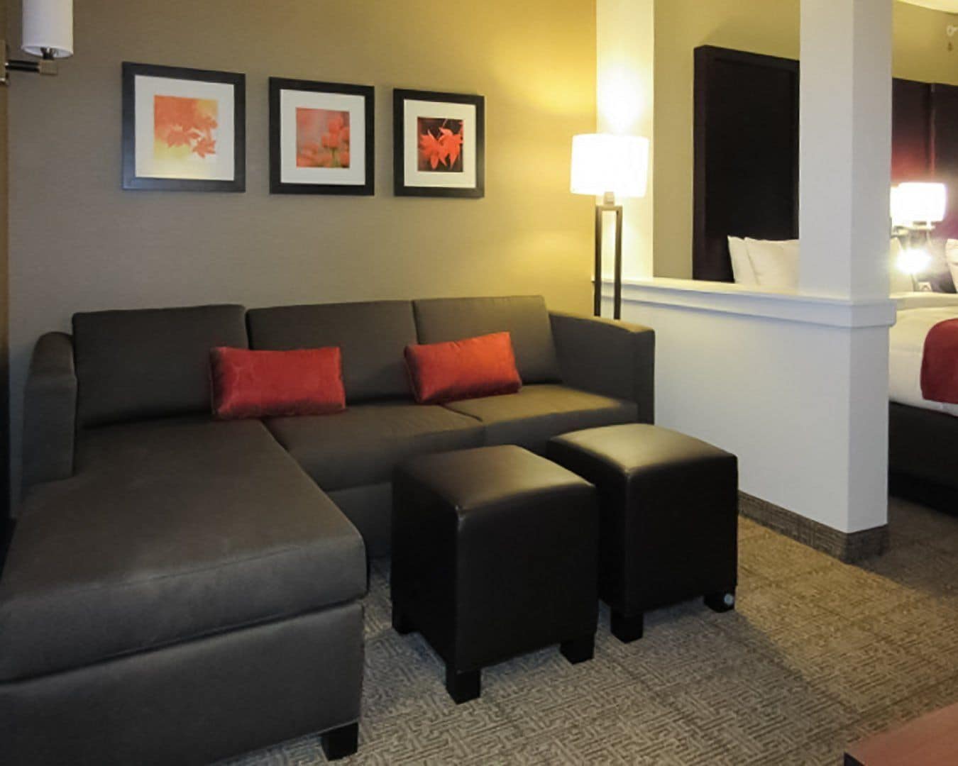 Un salón moderno con un sofá seccional gris, dos cojines rojos y un pequeño banquillo negro. Hay tres cuadros enmarcados en la pared y una lámpara de pie a la derecha.