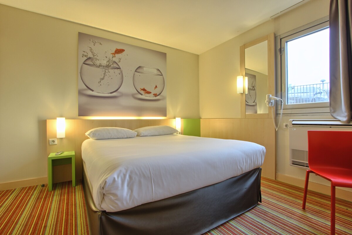 Ein Hotelzimmer mit einem Bett, einem grünen Nachttisch, einem roten Sessel, einem großen Fenster und einem Wandbild mit Fischen im Wasser.