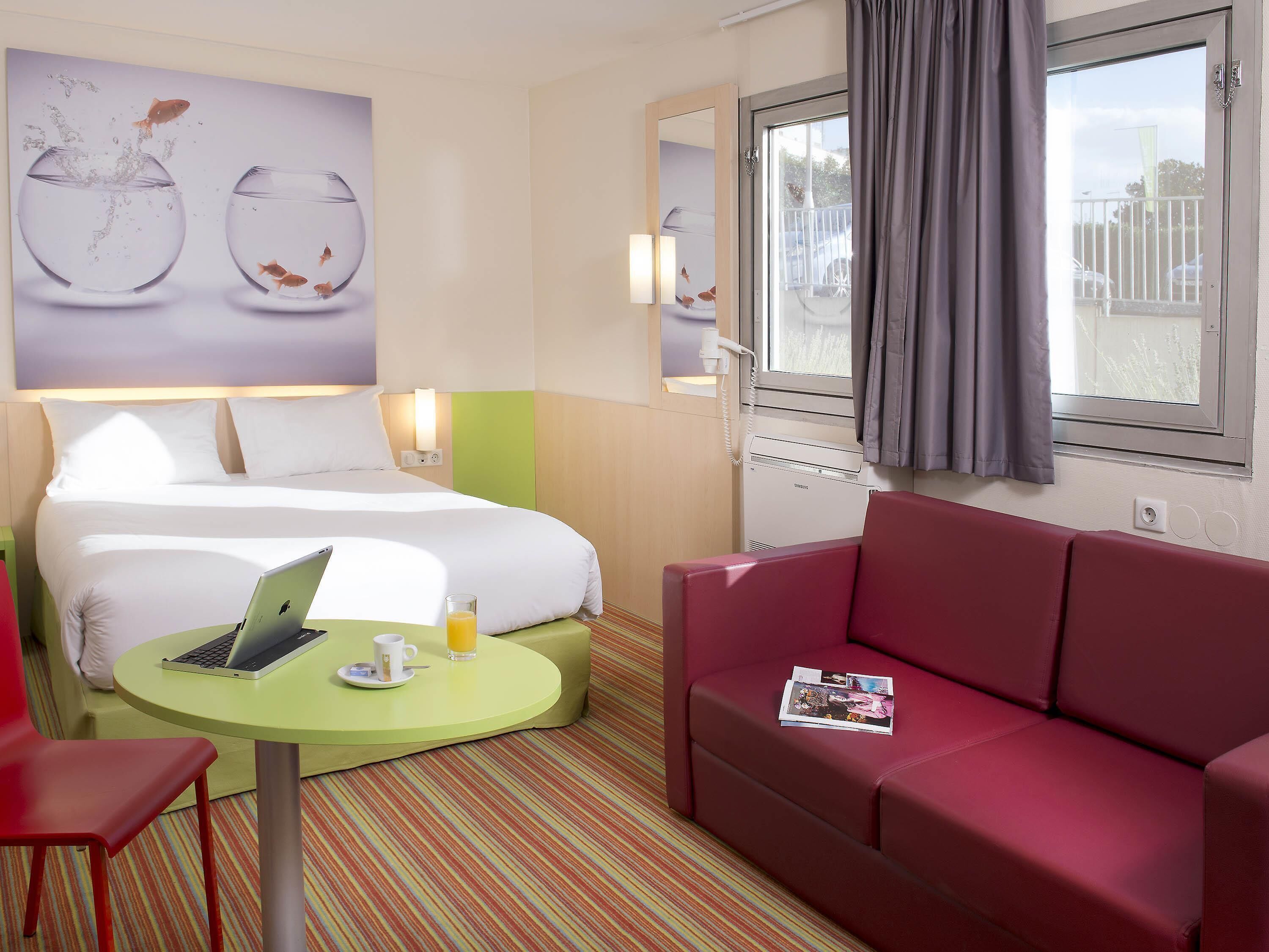 A hotel room with a bed, a green table with a laptop, a red couch, and a window with a view of the outside.