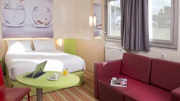 A hotel room with a bed, a green table with a laptop, a red couch, and a window with a view of the outside.