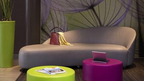 A modern living room with a grey sofa, a green and a pink ottoman, and a laptop on one of the ottomans. - ibis Styles Paris Roissy CDG (Roissy-en-France)
