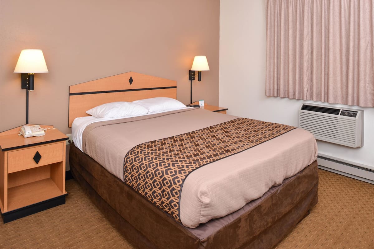 Quarto de hotel com cama, duas mesas de cabeceira, luminária, telefone e aparelho de ar condicionado.