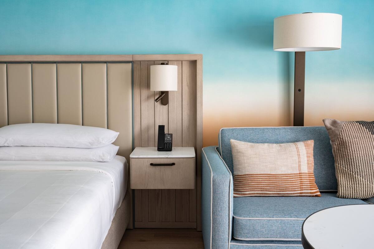 A hotel room with a bed, a nightstand with a lamp, a sofa with cushions, and a wall-mounted phone.
