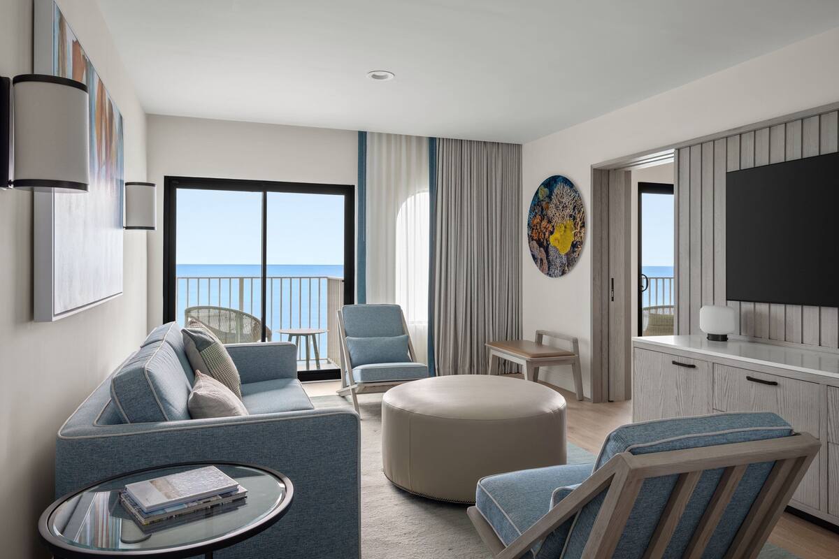 A modern hotel room with a balcony, a sofa, a round ottoman, a small table, and a TV.