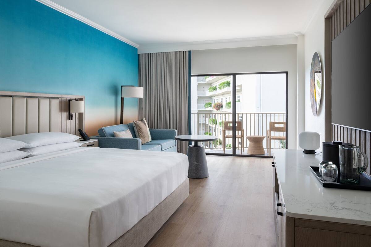 A hotel room with a large bed, a sofa, a small table, and a balcony with a view of the building.
