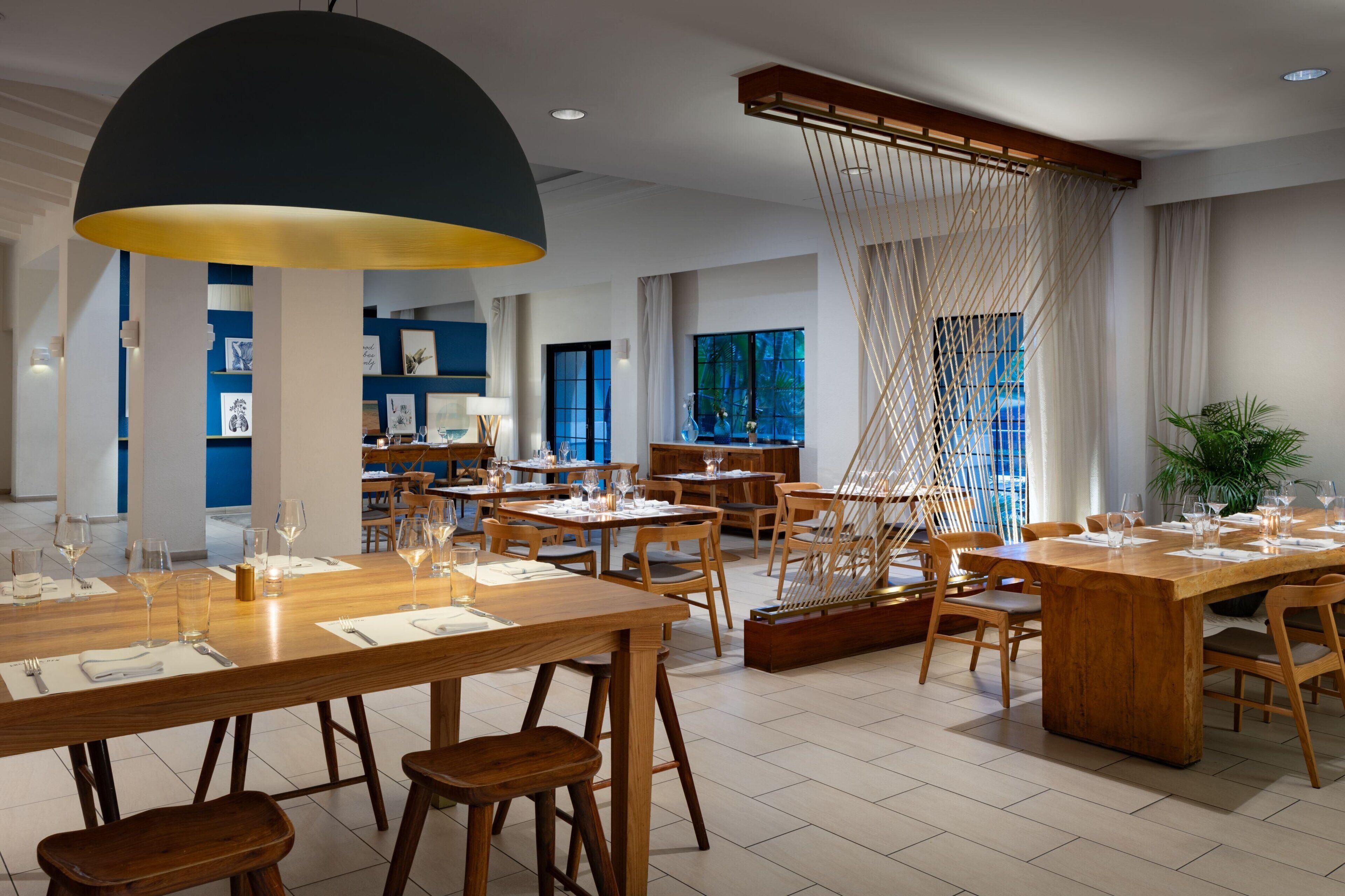 A modern dining area with wooden tables and chairs, a large black pendant light, and a view of the outdoors through large windows.