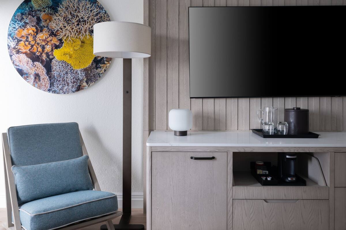 A modern living room with a blue armchair, a round wall art piece, a floor lamp, a flat-screen TV, and a coffee station.