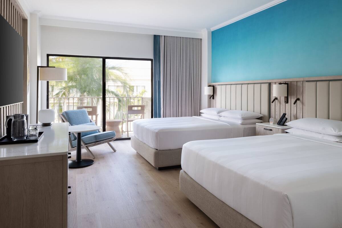 A hotel room with two beds, a desk with a lamp, a chair, and a view of the outdoors.